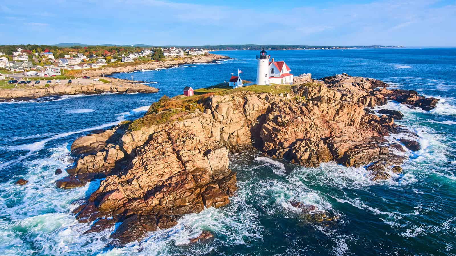 Waves crash over rocks aerial over Maine island with lighthouse and view of homes