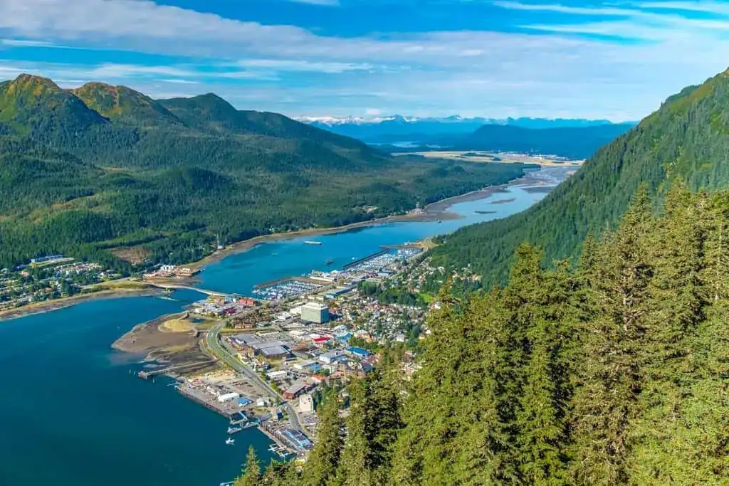 AK - An Alaska coastal town between green mountains and a winding river, with a marina and distant snow-capped peaks.